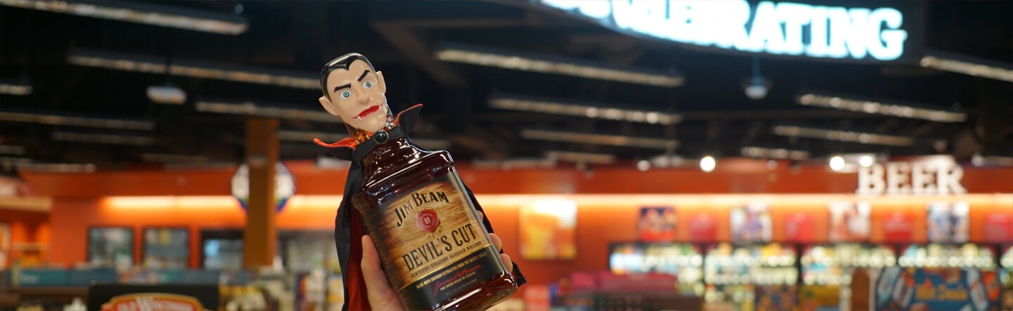 Image of someone holding a Jim Beam Devil's Cut with a vampire head and a cape on the bottle, and #AlwaysBeCelebrating sign behind.