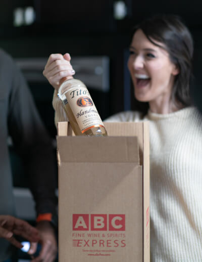 Host pulling out a Tito's Handmade Vodka bottle from an ABC Fine Wine & Spirits Express box.